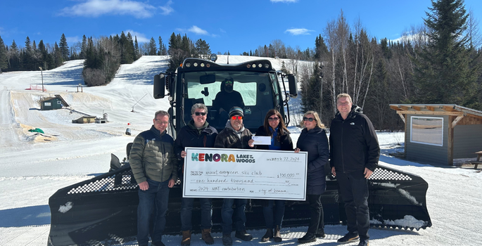 Group of people standing by a ski hill groomer on a ski hill holding a donation cheque